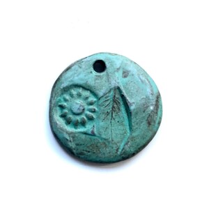 May include: A round, turquoise-colored pendant with a hole at the top. The pendant features embossed designs of a spiral and a leaf. The surface has a textured, aged appearance, suggesting a handcrafted or vintage style.