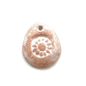 May include: A handmade, teardrop-shaped pendant with a sunburst design. The pendant is made of terracotta clay and has a hole at the top for stringing. The pendant is a light brown color with white accents.