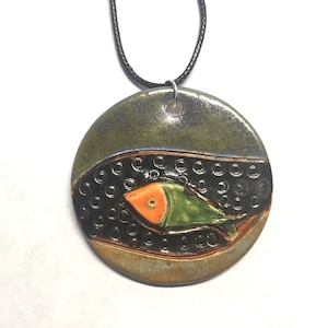 May include: A round ceramic pendant necklace with a green, orange, and black fish design. The pendant is on a black cord.