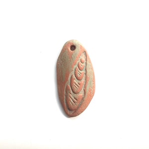 May include: A small, oval-shaped pendant made of clay with a carved design of a seashell. The pendant is a light brown color with a hint of green.