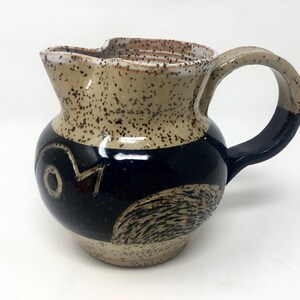 May include: A brown and black ceramic pitcher with a speckled glaze and a stylized bird design. The pitcher has a wide mouth and a handle.