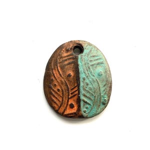 May include: Oval-shaped pendant with a hole at the top. The pendant is divided into two sections: one side is brown with etched patterns, and the other is turquoise with similar designs. The pendant has a rustic, handcrafted appearance.