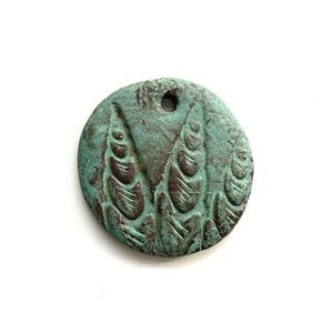 May include: A round, weathered metal pendant with a turquoise and brown patina. The pendant features a raised, stylized plant design and a hole for stringing. The overall aesthetic is rustic and antique.