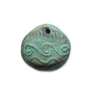 May include: A round, teal-colored pendant with a weathered, oxidized finish. The surface features a dark, embossed wave pattern across the center and a small hole at the top for suspension. The edges are irregular and rounded, showing dark brown tones.