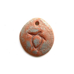 May include: A round, terracotta-colored pendant with a silver-colored turtle design. The pendant has a hole at the top for hanging.