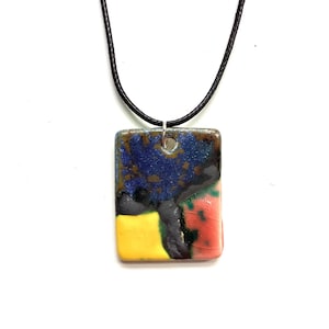 Puede incluir: Un collar con un colgante de cerámica rectangular de colores. El colgante presenta un diseño azul, amarillo, rojo y negro. El collar está hecho con un cordón negro.