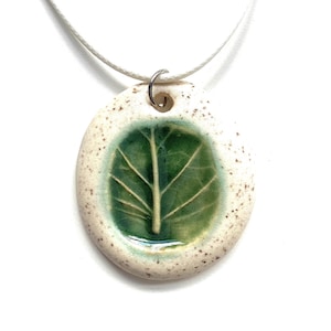 May include: A white ceramic pendant necklace featuring a green leaf design. The pendant is oval shaped and has a speckled white glaze. The leaf is a dark green colour and has a detailed vein pattern.