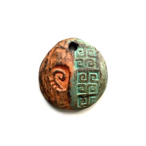 May include: A handcrafted, round pendant with a hole at the top. The pendant is divided into two sections: one side is orange with a spiral design, and the other is green with a geometric pattern. The pendant has a rustic, aged appearance.