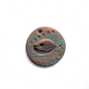 May include: A round, terracotta-colored ceramic ornament with a blue-green glaze. The ornament features a stylized fish design with a raised, textured surface.