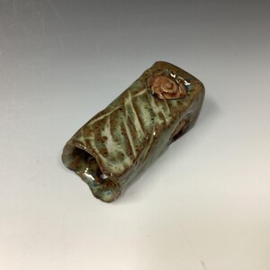 May include: A small, hand-crafted ceramic vase with a green and brown glaze. The vase has a textured surface and a small, pink flower detail on the top.