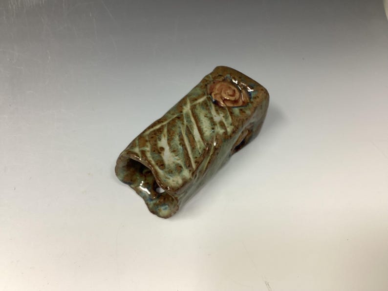 May include: A small, hand-crafted ceramic vase with a green and brown glaze. The vase has a textured surface and a small, pink flower detail on the top.