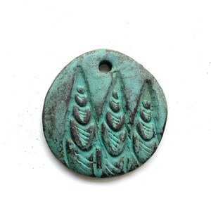 May include: A round, teal-colored pendant with a textured surface and a hole at the top. The pendant features a raised design of three stylized, vertical shapes. The overall aesthetic is rustic and handcrafted.