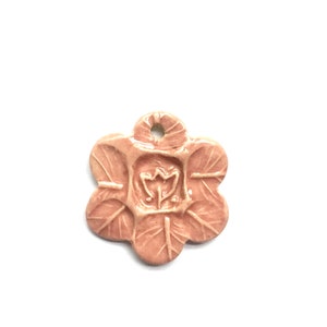 May include: A pale pink ceramic charm shaped like a flower with five petals. The center of the flower has a raised design of a stylized flower.