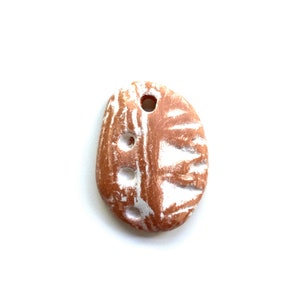 May include: A small, oval-shaped ceramic pendant with a white glaze and brown clay showing through. The pendant has a hole at the top for hanging and a design of white lines and circles.
