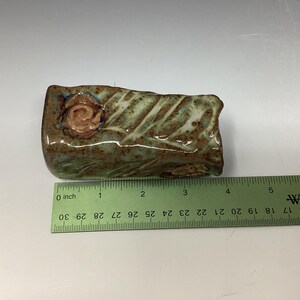 May include: A small, rectangular ceramic sculpture with a green and brown glaze. The sculpture has a raised, pink rose design on one side. The sculpture is approximately 3 inches long.