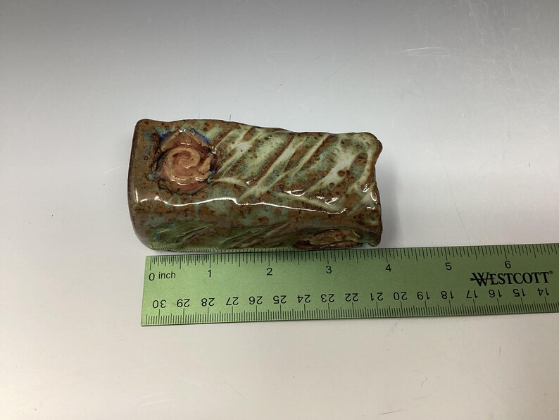 May include: A small, rectangular ceramic sculpture with a green and brown glaze. The sculpture has a raised, pink rose design on one side. The sculpture is approximately 3 inches long.