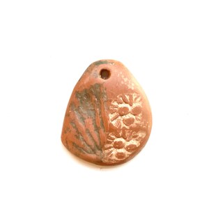 May include: A terracotta pendant with a hole at the top. One side has a carved design of a flower with a center and petals. The other side has a carved design of a leaf.