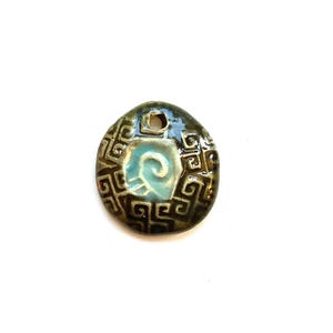 May include: A ceramic pendant with a dark brown border and a light blue center. The pendant has a spiral design in the center and a geometric pattern around the edges. It has a hole at the top for hanging.