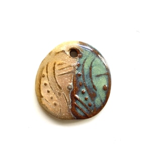 May include: A round ceramic pendant featuring a split design with a tan, textured left side and a glossy, teal-green right side. The surface is decorated with embossed dots and lines, and a small circular hole is positioned at the top center.