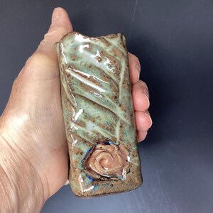 May include: A small, green and brown ceramic vase with a textured surface and a raised, pink rose design.