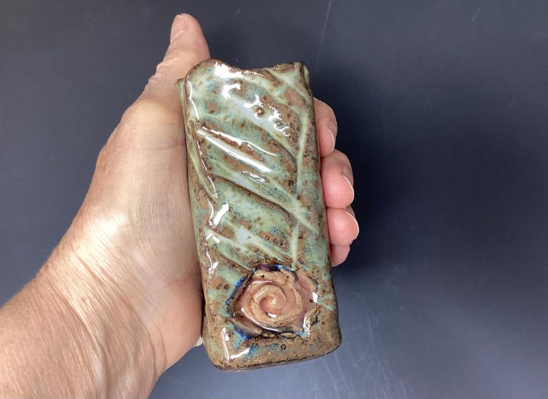 May include: A small, green and brown ceramic vase with a textured surface and a raised, pink rose design.
