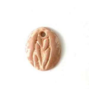 May include: A round, light brown ceramic pendant with a raised design of a stylized flower. The pendant has a hole at the top for hanging.