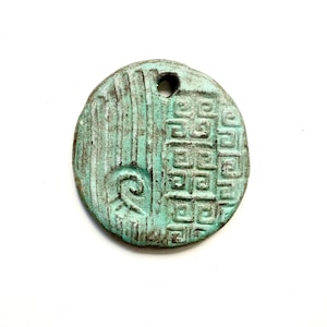 May include: A circular pendant with a weathered teal and bronze patina finish. The surface features a split design with vertical lines and a spiral on the left, and a repeating geometric square pattern on the right. A small hole is at the top.