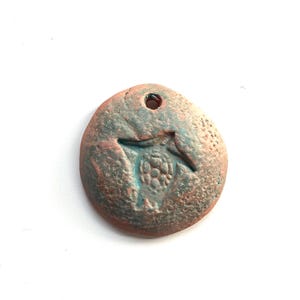 May include: A round, copper-colored pendant with a textured surface and a small hole at the top. The pendant has a design of a stylized bird with a raised, textured body.
