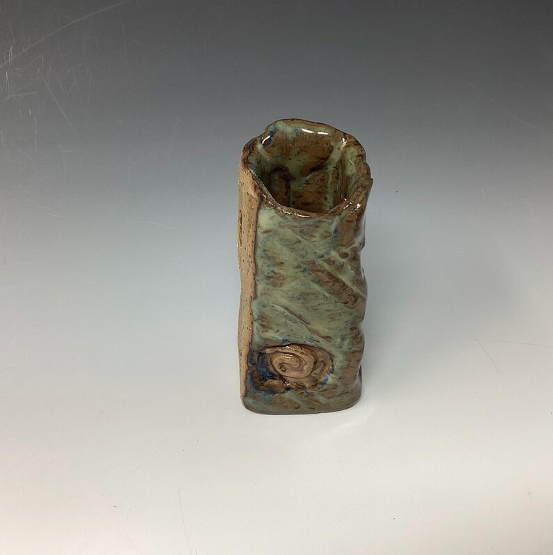 May include: A small, handmade ceramic vase with a green and brown glaze. The vase has a textured surface and a unique, irregular shape.
