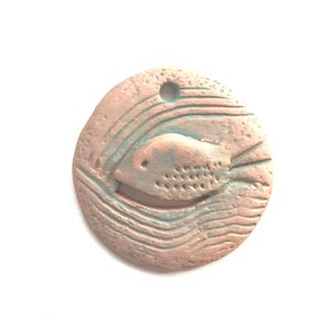May include: A round, ceramic pendant with a light brown and blue glaze. The pendant features a raised, detailed design of a fish swimming in wavy lines.