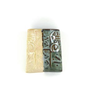 May include: A rectangular ceramic object with two distinct sections. One side is a light beige with embossed swirls, the other is a teal green with embossed geometric shapes. The object is likely a decorative piece or a small container.