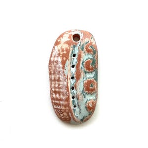 May include: A handcrafted, oblong pendant with a hole at the top. The pendant features a blend of red-orange and light blue-green hues, with a textured surface and drilled holes. The design includes a series of small, dark dots.