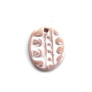May include: An oval-shaped clay pendant with a textured, light brown surface and white painted accents. The piece features a central vertical line with dots and geometric patterns on either side, and a small hole at the top for hanging.