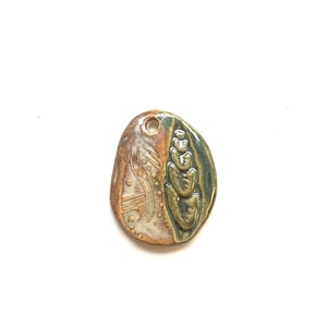 May include: An oval ceramic pendant featuring a glossy finish with a split design. The left side displays a textured pattern of dots and lines in shades of tan and brown, while the right side features a raised, dark green organic leaf-like motif.