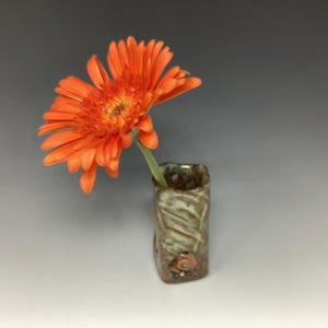 May include: A small, green ceramic vase with a textured surface and a decorative shell. A single orange flower is in the vase.