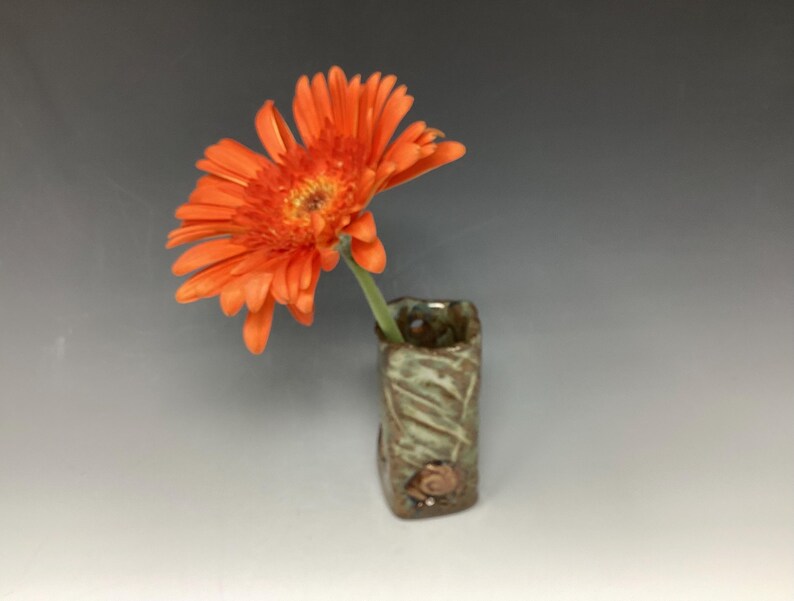 May include: A small, green ceramic vase with a textured surface and a decorative shell. A single orange flower is in the vase.