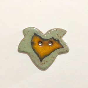 May include: A ceramic button with a green and brown glaze. The button has a unique shape and two holes. The center of the button is a bright orange color.