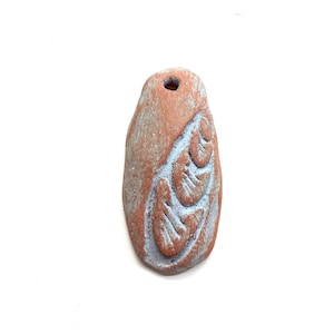 May include: A terracotta pendant with a carved design of a leaf. The pendant is a light brown color with a slightly textured surface.