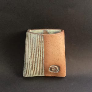 Puede incluir: Un florero de pared de cerámica con parte posterior plana y forma rectangular cónica. El lado izquierdo presenta líneas verticales grabadas en esmalte verde menta pálido, mientras que el derecho es de arcilla beige moteada con un detalle en espiral.