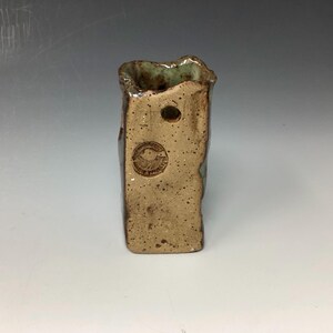 May include: A small, rectangular ceramic vase with a rough, textured surface. The vase is glazed in shades of brown and green, with a small hole near the top. The bottom of the vase is stamped with a circular logo.