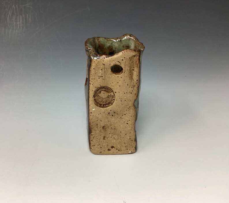 May include: A small, rectangular ceramic vase with a rough, textured surface. The vase is glazed in shades of brown and green, with a small hole near the top. The bottom of the vase is stamped with a circular logo.