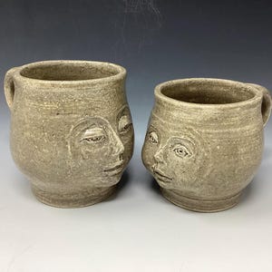 Puede incluir: Dos tazas de cerámica con caras esculpidas en la parte delantera. Las tazas son de color marrón claro con una superficie texturizada. Las caras se miran.