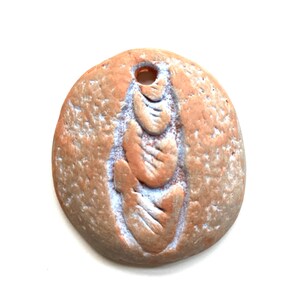 May include: A round, terracotta-colored ceramic ornament with a raised, white, shell-like design. The ornament has a small hole at the top for hanging.