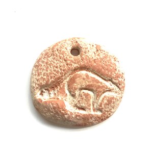 May include: A circular, reddish-brown pendant featuring a carved animal design. The pendant has a textured surface and a hole for threading. The animal carving is a lighter shade of brown. The pendant is likely made of stone.