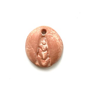 May include: A round, coral-colored pendant with a textured surface and a central, vertical design. The pendant has a hole at the top for stringing. The design is raised and detailed, adding depth to the piece. The pendant is set against a white background.