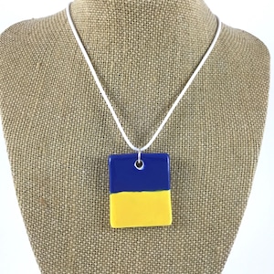 May include: A square pendant necklace featuring a blue and yellow colour block design. The pendant hangs from a white cord. The necklace is displayed on a burlap-textured surface.