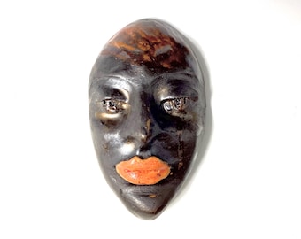 Könnte beinhalten: Eine Keramikmaske mit schwarzem Gesicht, braunen und orangefarbenen Akzenten und orangefarbenen Lippen.