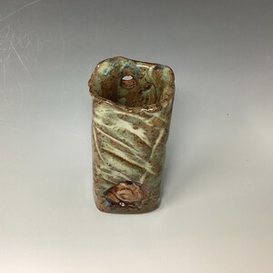 May include: A small, rectangular ceramic vase with a textured, green and brown glaze. The vase has a hole on the side and a small, raised design on the front.