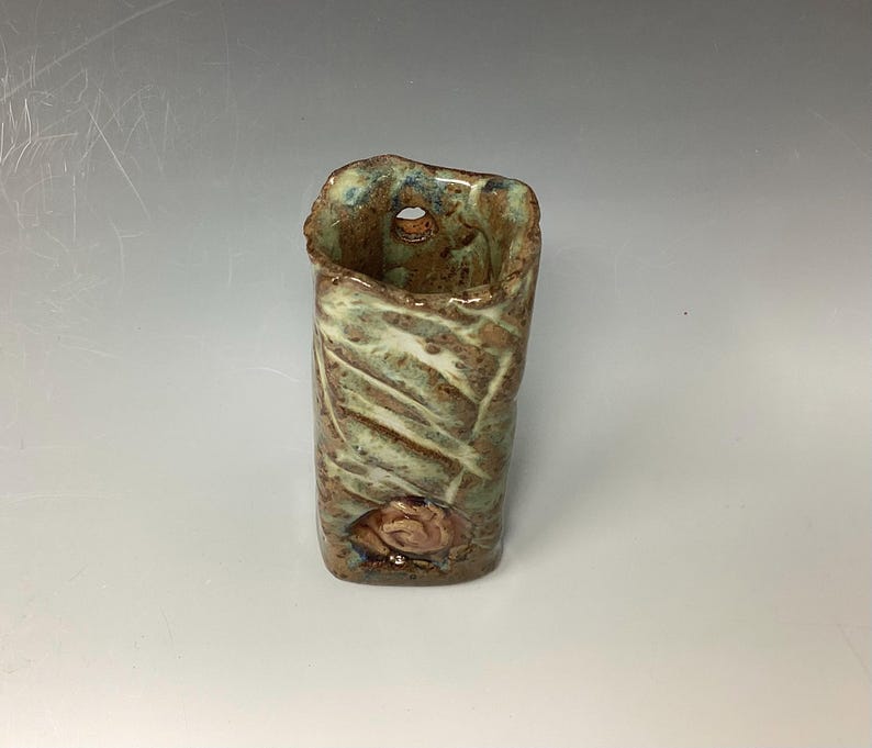 May include: A small, rectangular ceramic vase with a textured, green and brown glaze. The vase has a hole on the side and a small, raised design on the front.