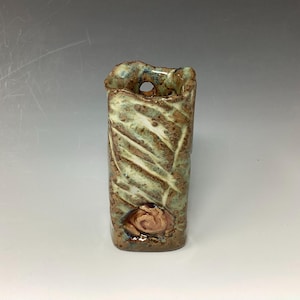 May include: A small, green and brown ceramic vase with a textured surface and a small hole near the top. The vase has a decorative design on the front.